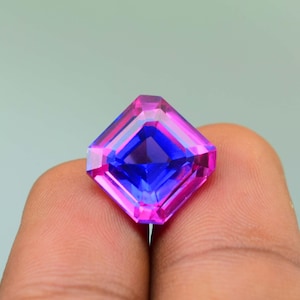 May include: A square-cut gemstone with a striking colour gradient. The gem transitions from a deep blue centre to a bright pink hue at the edges. The gem is held between two fingers.