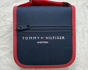 May include: A square navy blue fabric case with a red trim and a top handle. The front features a horizontal zipper and the white embroidered text Tommy Hilfiger watches with the brand's signature red, white, and blue flag logo.