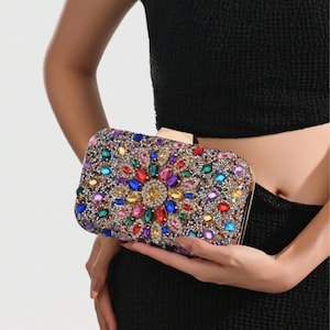 Puede incluir: Clutch rectangular cubierta de pedrería colorida. El diseño presenta un patrón floral central con gemas rojas, azules, amarillas y verdes. El clutch tiene un cierre y marco dorados.