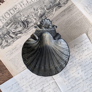 May include: A dark metallic scallop shell dish featuring a decorative fan-shaped top edge and deep vertical ridges. The surface has a weathered, tarnished finish with subtle golden highlights along the center and edges of the shell-shaped bowl.