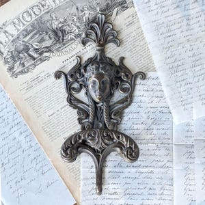 May include: A dark metallic wall hook featuring a central sculpted human face surrounded by intricate scrollwork and foliage patterns. The hook has a vertical orientation with a decorative finial at the top and a curved hook at the bottom.