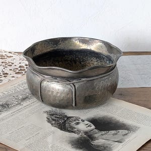 May include: A silver-toned metal bowl with a scalloped rim and a hammered texture on the exterior. The bowl features a decorative rope-like trim encircling the upper section, with vertical rope accents extending down the sides of the rounded body.