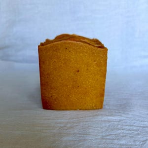 May include: A rectangular bar of soap with a textured, golden-orange surface. The soap has a slightly uneven, rustic top edge and a dense, granular appearance throughout its body, showcasing a consistent warm, earthy tone across its front face.