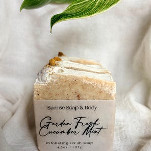 May include: A bar of exfoliating scrub soap with a light beige color and a textured surface. The soap is topped with a white swirl design and small yellow granules. The label reads "Garden Fresh Cucumber Mint" and "Sunrise Soap & Body".
