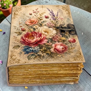 May include: A rectangular journal with a beige cover featuring a colorful floral bouquet of pink, red, and white flowers. The cover has ornate gold-toned filigree borders and a dark metal latch. The pages have a textured, aged yellow appearance.