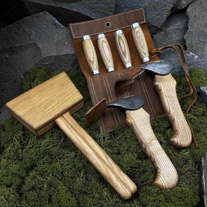 Könnte beinhalten: Ein Holzhammer und ein Satz von vier Meißeln mit Holzgriffen und Metallklingen. Die Werkzeuge werden auf einem Bett aus grünem Moos präsentiert, mit einer Leder-Werkzeugrolle im Hintergrund. Der Hammer und die Meißel sind neu.