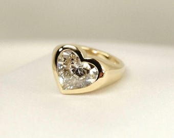 Op de afbeelding: Een gouden ring met een hartvormige zetting die een enkele, gefacetteerde, heldere edelsteen bevat. De ringband is glad en gepolijst, met een afgerond profiel dat naar achteren toe taps toeloopt, wat zorgt voor een strakke, reflecterende afwerking.