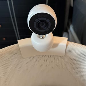 May include: A white Harbor baby monitor camera is mounted on a white mesh surface. The camera has a black lens and the word "Harbor" is printed on the front. The camera is attached to a white base.