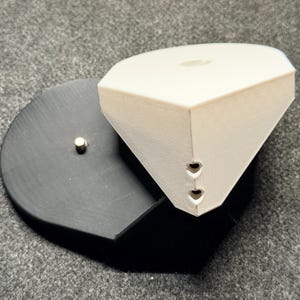 May include: A 3D printed object featuring a white, multi-faceted geometric top section with two small heart-shaped cutouts on the front edge, mounted onto a flat, black, heart-shaped base that includes a single small metallic stud component.
