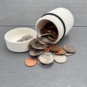 May include: A white textured coin holder with a black band, partially tipped over, spilling out various coins. The holder's lid is open, revealing more coins. The coins are scattered on a gray surface, including pennies, dimes, quarters, and nickels.