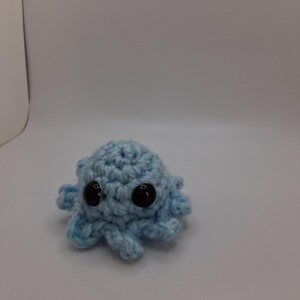May include: A small, light blue crocheted octopus with large, black safety eyes. The handmade plush toy has eight curled tentacles and is set against a white background. The octopus is a cute, handmade item.