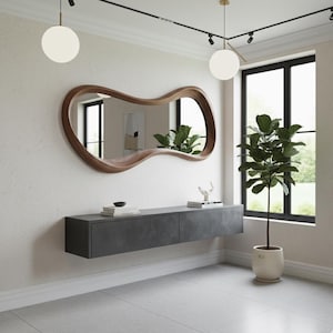 Op de afbeelding: Een modern interieur met een grote, uniek gevormde spiegel met houten frame. Daaronder een zwevend dressoir in donkergrijs. Een potplant en twee bolvormige hanglampen zijn ook zichtbaar, wat een strakke sfeer cre&euml;ert.