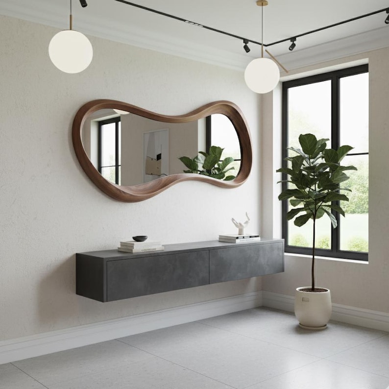 Op de afbeelding: Een modern interieur met een grote, uniek gevormde spiegel met houten frame. Daaronder een zwevend dressoir in donkergrijs. Een potplant en twee bolvormige hanglampen zijn ook zichtbaar, wat een strakke sfeer cre&euml;ert.