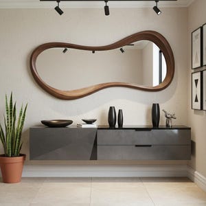 Op de afbeelding: Een moderne woonkameropstelling met een grote, uniek gevormde houten spiegel. Onder de spiegel staat een grijze zwevende kast met twee lades. Decoratieve zwarte vazen en een klein beeldje staan op de kast. Een plant in een pot staat links.