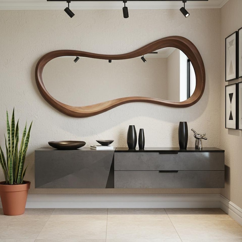 Op de afbeelding: Een moderne woonkameropstelling met een grote, uniek gevormde houten spiegel. Onder de spiegel staat een grijze zwevende kast met twee lades. Decoratieve zwarte vazen en een klein beeldje staan op de kast. Een plant in een pot staat links.