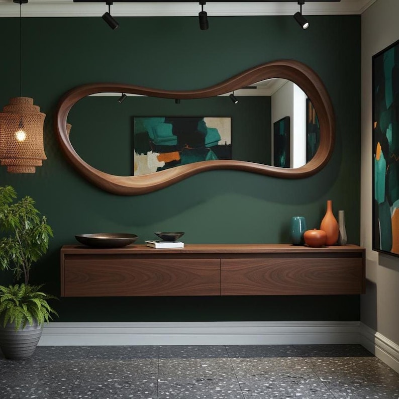 Op de afbeelding: Een modern interieur met een grote, organisch gevormde houten spiegel. Onder de spiegel een zwevende houten console met decoratieve items, waaronder vazen en een kom. De muur is diepgroen geschilderd en de vloer is grijs gespikkeld.