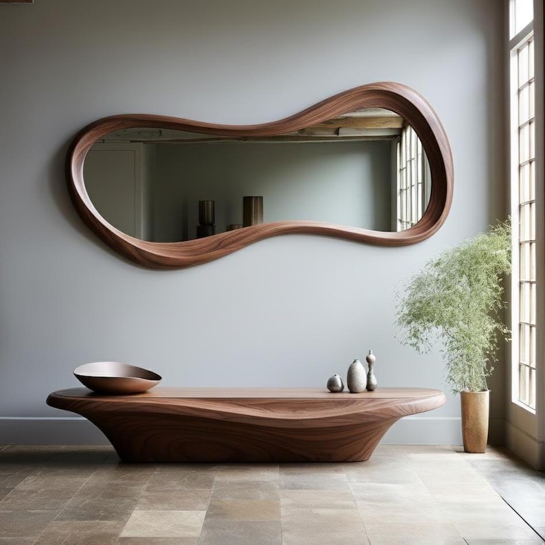 Op de afbeelding: Een grote, organisch gevormde houten spiegel hangt boven een bijpassende houten salontafel. De lijst van de spiegel en de tafel delen een rijke, donkerbruine kleur. Een decoratieve kom en kleine vazen sieren de tafel en vullen de minimalistische esthetiek van de kamer aan.