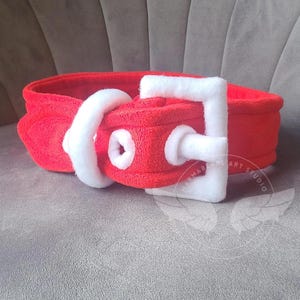 May include: A plush red belt with a white buckle and accents. The belt is made of a soft, textured material and features a classic buckle design. The belt is designed to be worn around the waist.