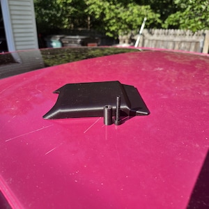 May include: A black, rectangular, low-profile plastic cover with a textured surface, accompanied by a small black cylindrical spacer and a single black metal bolt with a threaded shaft and a hexagonal head, all resting on a smooth magenta surface.
