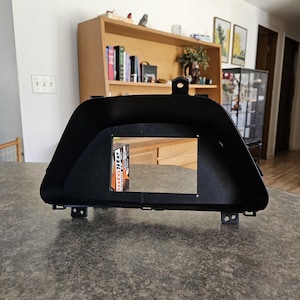 Può includere: Una cornice nera del cruscotto con un'apertura rettangolare. La cornice ha una superficie strutturata e linguette di montaggio. All'interno dell'apertura è visibile una scatola di candele "AutoLite Iridium XP".