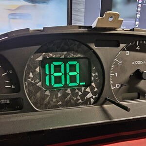 May include: A black automotive instrument cluster featuring a digital display showing the number 188 MPH in bright green. The display is surrounded by a dark gray geometric pattern. To the right is an analog tachometer with white markings.