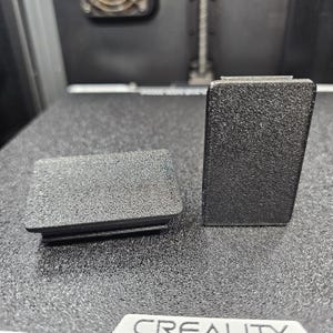 May include: Two black, rectangular plastic plugs with a textured surface. One plug is lying flat, while the other stands upright. The plugs appear to be components of a larger device or machine.