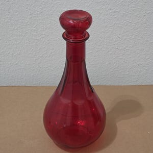 May include: A ruby red glass decanter with a round stopper. The decanter has a bulbous body and a long neck. The stopper is also round. The glass has a glossy finish.