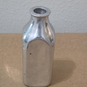 May include: A silver-coloured metal bottle with a square-shaped body and a rounded neck opening. The surface of the bottle has a smooth, reflective finish with subtle variations in tone and a few small dark spots visible on the front side.