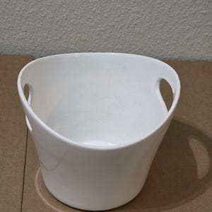 Peut inclure: Un seau en céramique blanche avec une finition lisse et brillante et une large ouverture sur le dessus. Le récipient présente deux découpes de poignées ovales intégrées sur les côtés opposés, avec un corps légèrement effilé.
