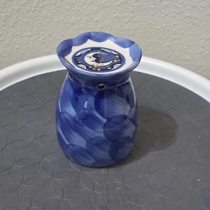 May include: A blue and white ceramic wax warmer featuring a hand-painted circular pattern on the body and a scalloped rim. The top dish displays a detailed illustration of a crescent moon surrounded by small yellow stars against a dark blue sky.