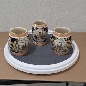 May include: Three ceramic beer steins featuring a dark blue body with a tan rim and base. Each stein displays a colourful illustration of the Munich skyline with the text München, surrounded by raised relief oak leaf and acorn decorations.