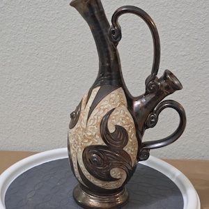 May include: A ceramic pitcher with a tall, slender neck and a secondary smaller spout. The body features a dark metallic brown glaze contrasted with cream-coloured, textured panels decorated with raised, swirling dark brown organic patterns.