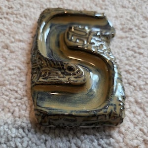 May include: A rectangular ceramic dish with a curved, flowing design. The dish features a glossy, two-tone glaze in shades of tan and blue. The edges are textured with a detailed pattern. The dish is likely handmade.