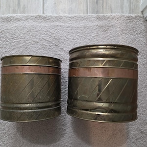 May include: Two cylindrical planters with a brass finish and copper bands. The planters have a textured, diagonal pattern. One planter is slightly smaller than the other. The planters are suitable for indoor or outdoor use.