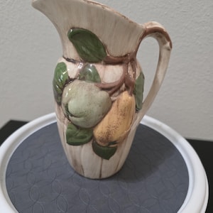 Puede incluir: Jarra de cerámica color crema con asa y boquilla. La jarra está decorada con hojas verdes y frutos de color verde claro y amarillo. La jarra está sobre una superficie blanca y gris.