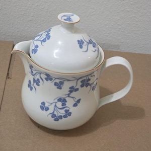 Op de afbeelding: Witte keramische theepot met deksel, versierd met een blauw bloemmotief. De theepot heeft een afgeronde body, een gebogen tuit en een handvat. Het deksel heeft een kleine knop. Het ontwerp omvat blauwe bloemtakken en een gouden rand.