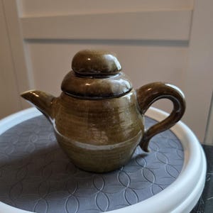 May include: A brown ceramic teapot with a rounded body, a curved handle, and a spout. The lid is a stacked design. The teapot has a glossy finish. The teapot is on a grey and white surface.