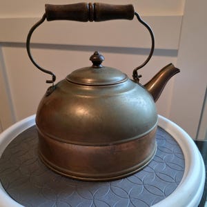 Pode incluir: Bule antigo de cobre e latão com uma pega de madeira. O bule tem um corpo arredondado, um bico e uma tampa com um pequeno botão. A pega é arqueada e presa às laterais. Está sobre uma superfície cinza e branca.