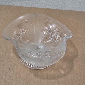 May include: A clear glass bowl with a ruffled, scalloped rim and a pedestal base. The exterior surface features a frosted, embossed pattern of pinecones and pine needles arranged in a repeating design around the sides of the circular vessel.
