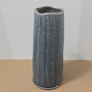 May include: A tall, cylindrical ceramic vase with a glossy, slate-blue glaze. The exterior surface features a repeating vertical pattern of impressed, fern-like leaf motifs that run from the base to the top rim, which has a slightly wavy edge.
