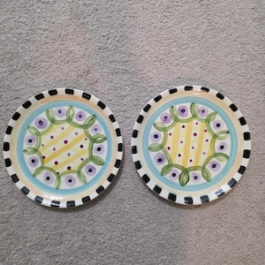 Könnte beinhalten: Zwei dekorative Keramikteller mit farbenfrohem Design. Jeder Teller hat eine gelb gestreifte Mitte, umgeben von einem hellblauen Ring mit lila floralen Akzenten. Der äußere Rand hat ein schwarz-weißes Schachbrettmuster.