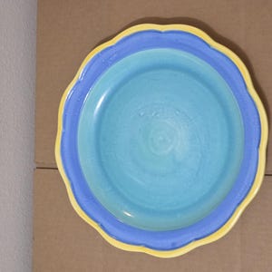 May include: A ceramic plate with a scalloped edge. The plate features a light blue center, a darker blue border, and a yellow rim. The plate is designed for serving food and adds a pop of color to any table setting.