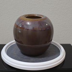May include: A spherical ceramic vase with a smooth, dark brown base that transitions into a glossy, mottled purple and amber glaze around the upper shoulder and rim. The vase features a wide, circular opening and a rounded, bulbous body shape.