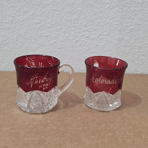 May include: Two small glass vessels with a deep red upper section and a clear, diamond-patterned base. The left vessel features a handle and the etched text "The Dells 1906", while the right vessel displays the etched word "Colorado" in script.