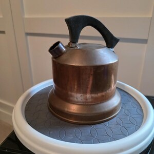 May include: A vintage copper tea kettle with a black handle and spout. The kettle sits on a gray patterned surface with a white rim. The kettle is a classic kitchenware item.
