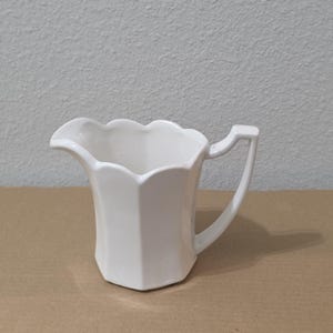 Puede incluir: Una jarra de leche de cerámica blanca sólida con un borde festoneado, un pico vertedor puntiagudo y un asa angular. El cuerpo de la jarra tiene una forma hexagonal con paneles verticales lisos que se estrechan hacia la base.