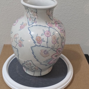May include: A white ceramic vase with a rounded body and a flared rim, featuring a repeating pattern of stylized pink, orange, and blue flowers and leaves with thin blue outlines across the entire exterior surface of the vessel.