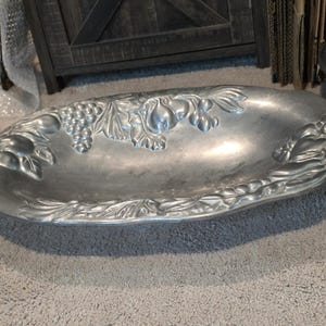 May include: A silver-colored, oval-shaped serving dish with embossed fruit and leaf designs. The dish features grapes, pears, and other fruits around the rim. The serving dish is made of metal and is suitable for serving food.