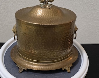 Carnicera antigua de latón martillado con tapa, patas y asas en forma de cabeza de león.