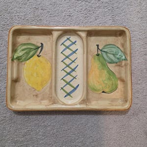 May include: A rectangular ceramic serving dish with three compartments. The dish features a yellow lemon and a green pear with leaves painted on the sides. The center section has a blue and green crisscross pattern.